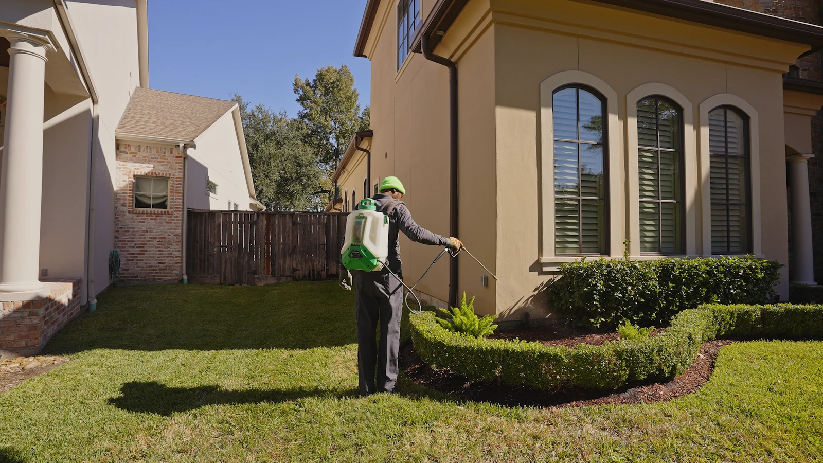 Pest control technician with a backpack sprayer treating the foundation of a suburban home